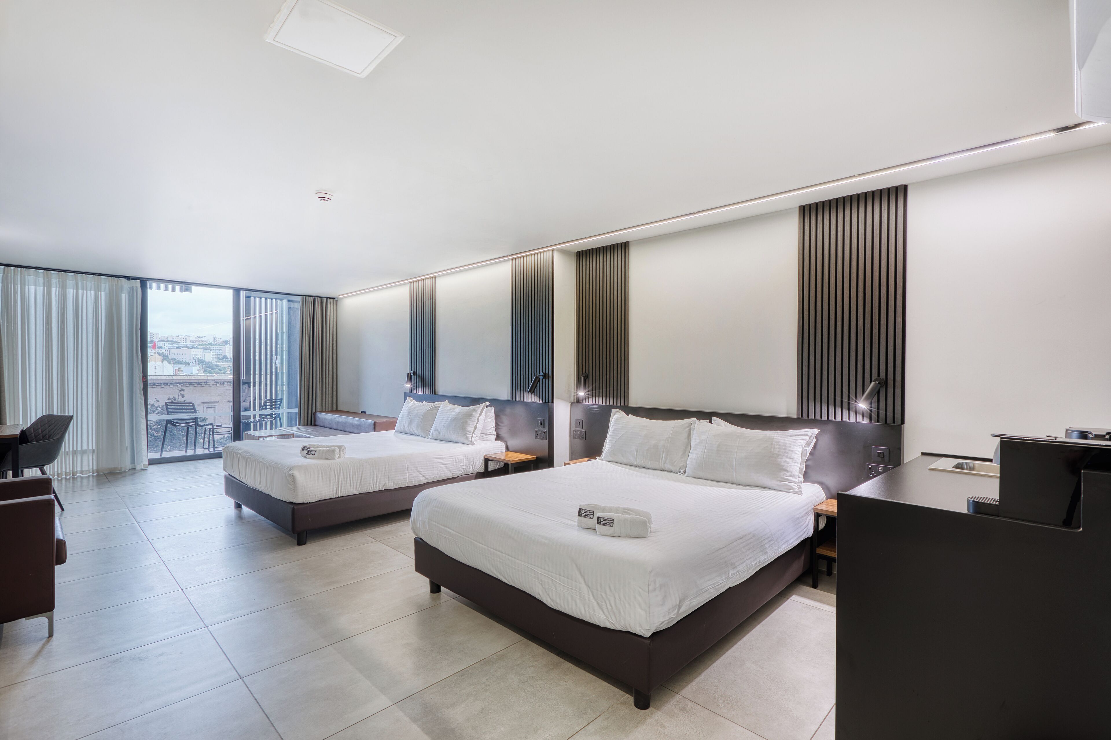 A modern hotel room with two beds, a desk, and a large window with a city view.