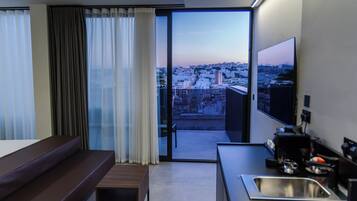 Una habitación de hotel moderna con balcón, cama, una mesita, televisor y una zona de cocina con lavabo.