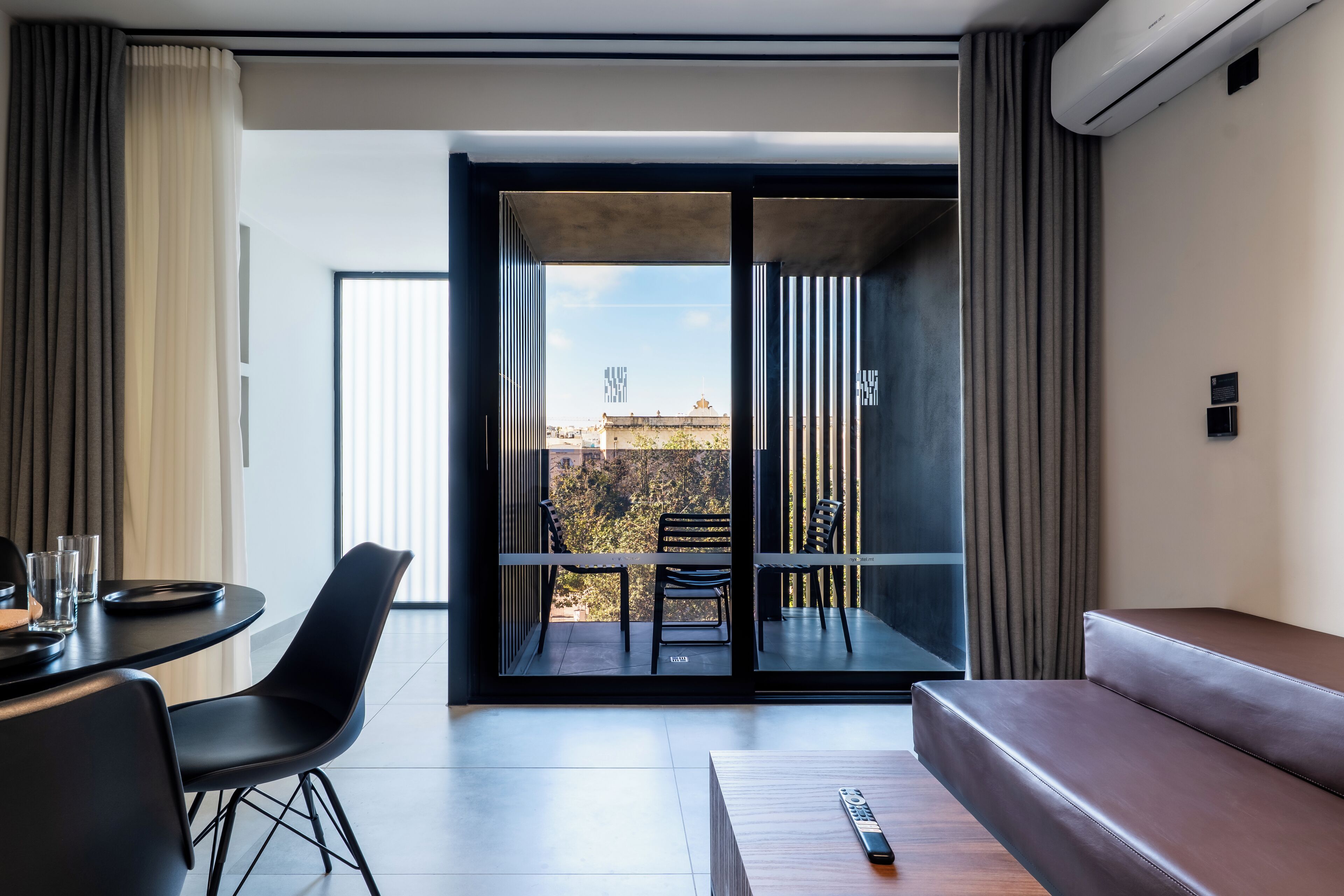 A modern hotel room with a balcony, a sofa, a dining table with chairs, and a remote control on the table.