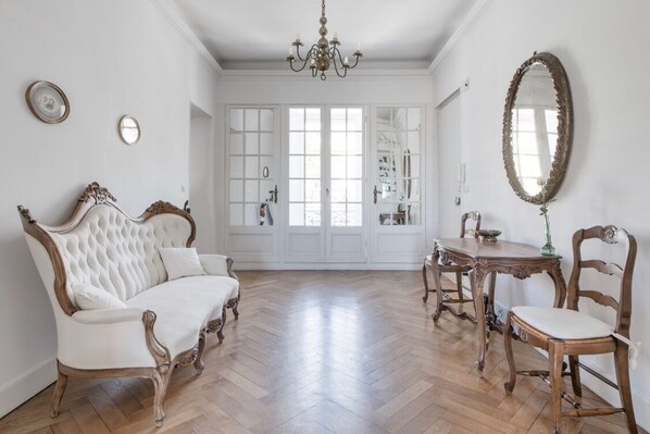 A spacious room with a white sofa, wooden dining table, and chairs, a chandelier, and a large mirror on the wall.