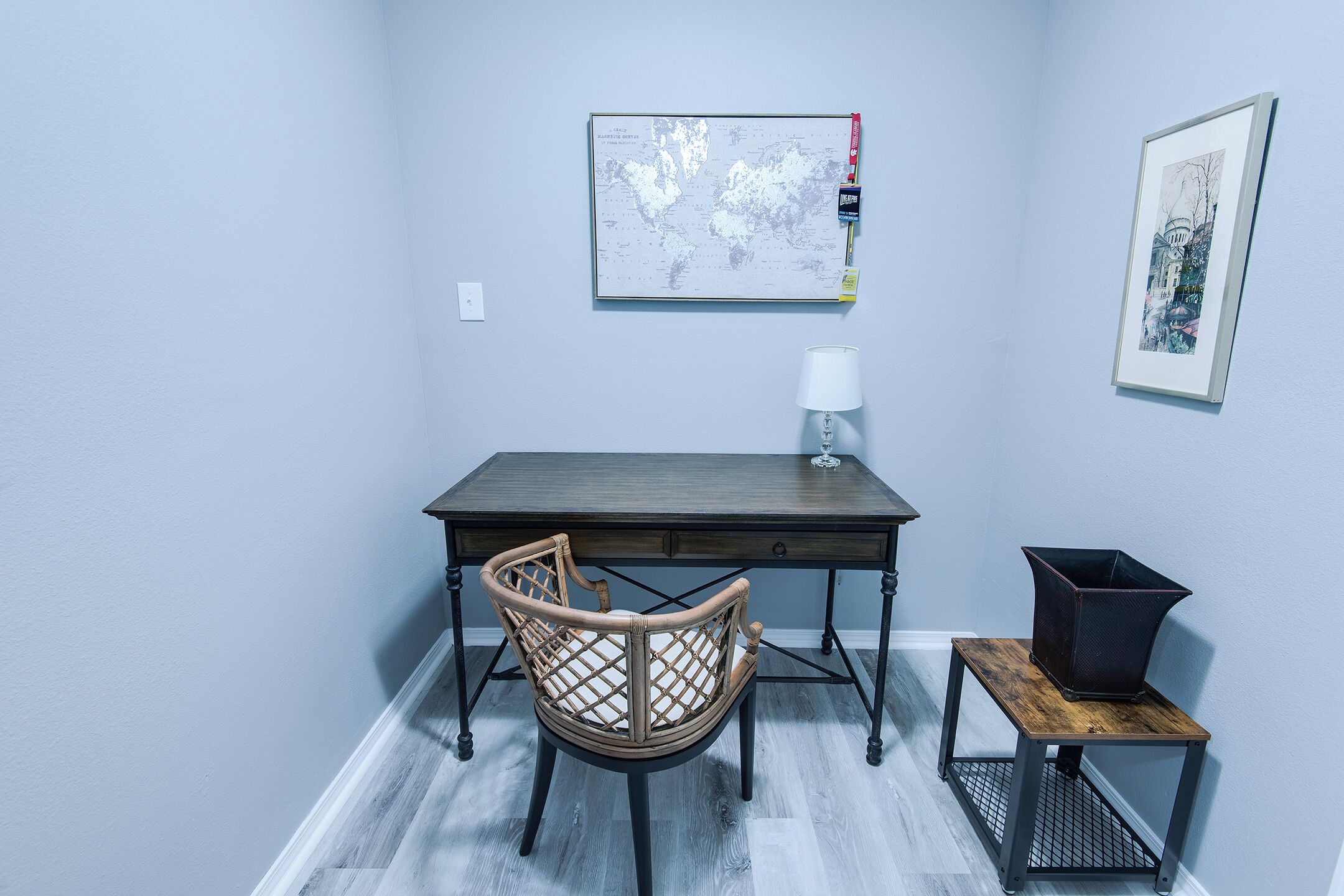 Une petite pièce, bien rangée, avec un bureau en bois, une chaise en osier, une petite table avec une lampe et une carte encadrée accrochée au mur.
