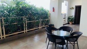 A covered outdoor seating area with a glass table and wicker chairs, surrounded by potted plants and a fire extinguisher on the wall.
