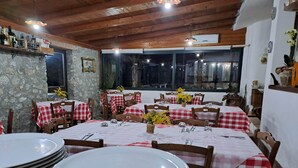 A restaurant with wooden ceilings and stone walls, featuring tables with red and white checkered tablecloths, wooden chairs, and a view of the night sky through large windows. -  Agriturismo Heaven (Ricadi)