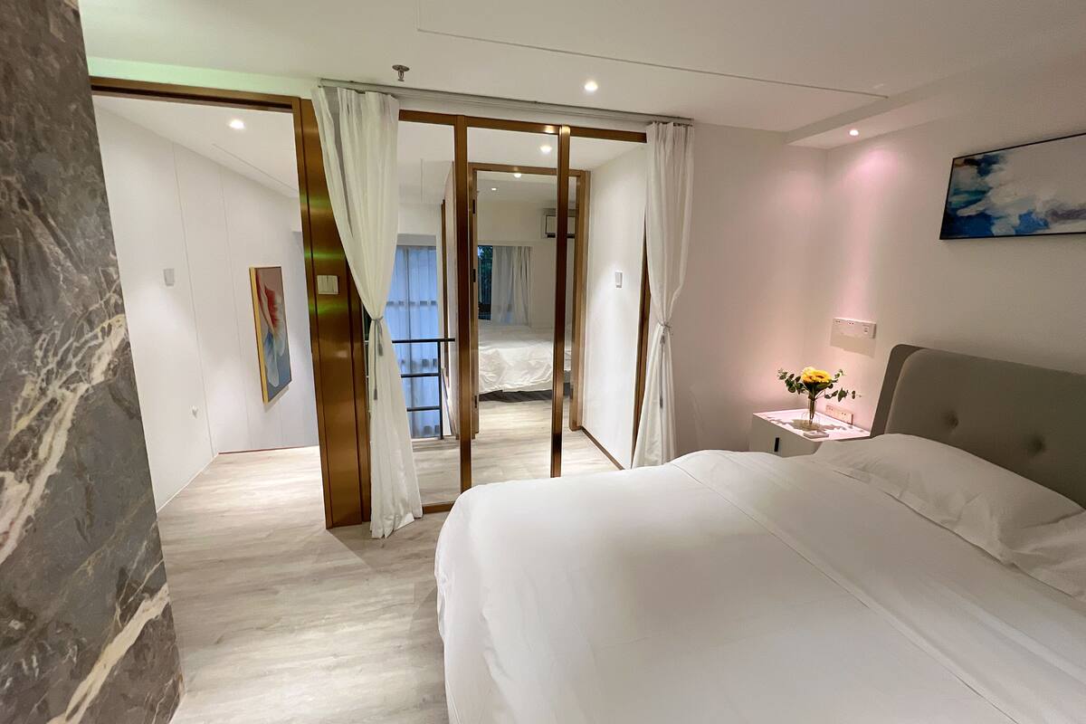 A hotel room with a bed, a nightstand, a painting, and a view of another room through a glass door.