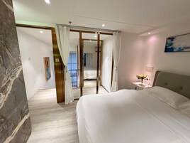 Habitación de hotel con cama, mesita de noche, un cuadro y vista a otra habitación a través de una puerta de cristal.