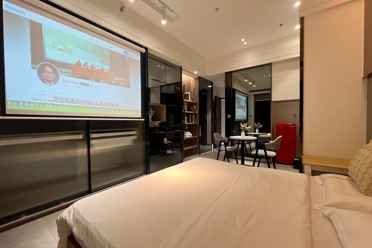A modern hotel room with a large bed, a desk with a chair, a small table with a vase, and a red cabinet.