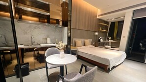 Une chambre d’hôtel moderne avec un lit, un bureau et une chaise. On y trouve une petite table sur laquelle est posé un vase de fleurs. La chambre est équipée d’un grand miroir et d’une fenêtre.