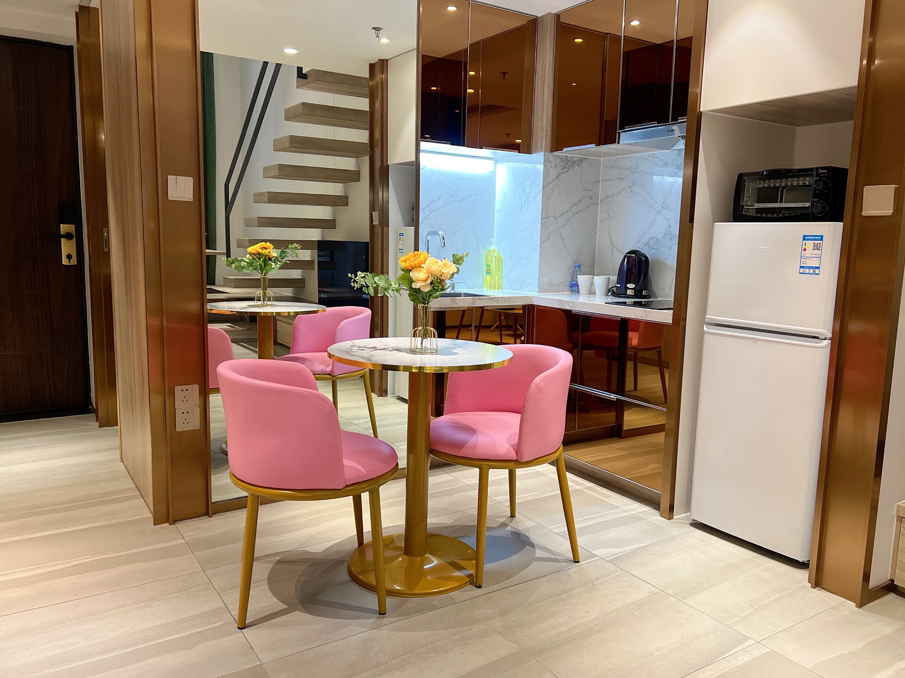 A modern kitchen with a built-in microwave, a refrigerator, and a coffee maker. There are pink chairs around a small table with a vase of flowers.