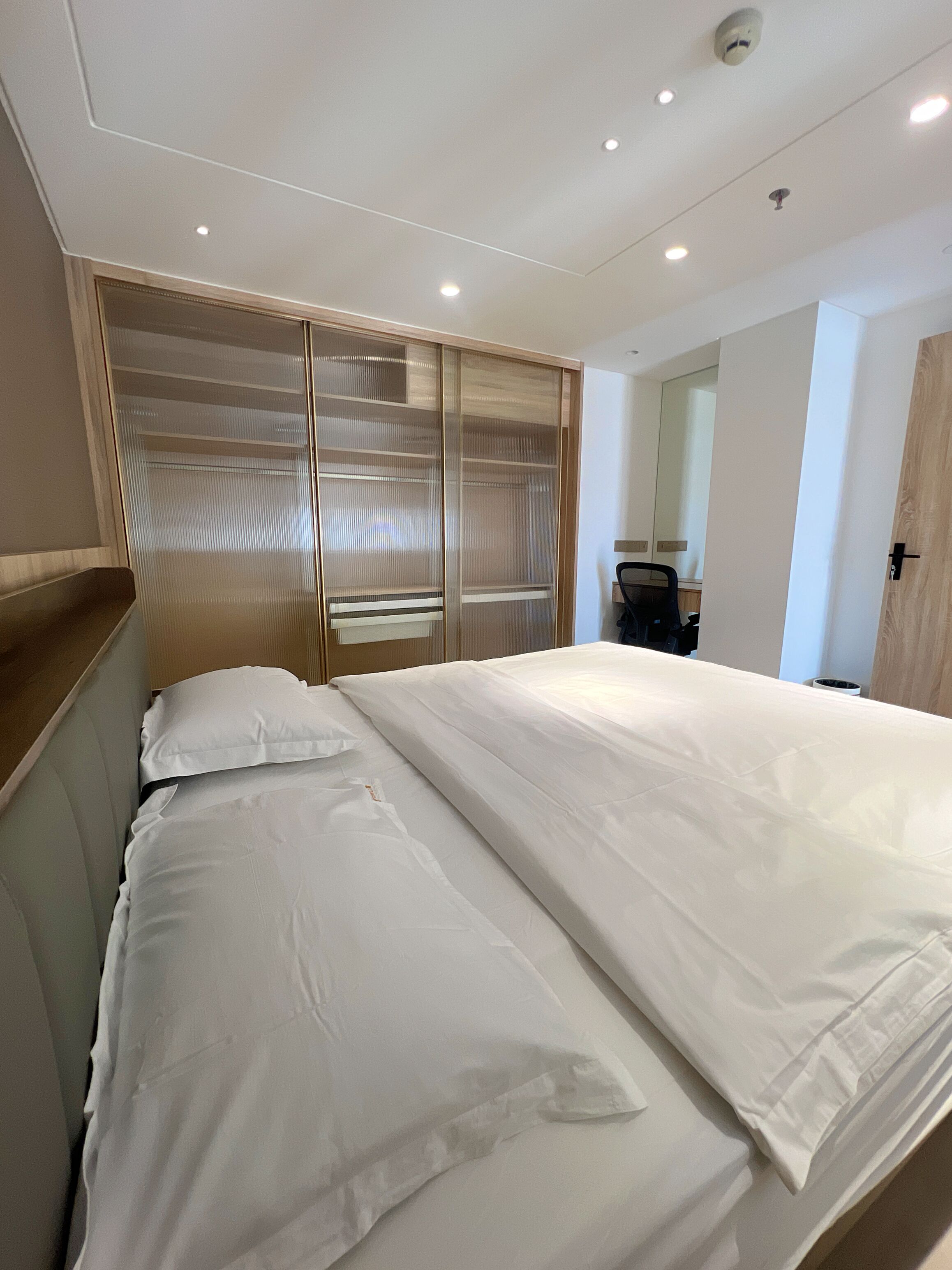 A modern hotel room with a large bed, a wooden headboard, a sliding glass door closet, and a desk with a chair.