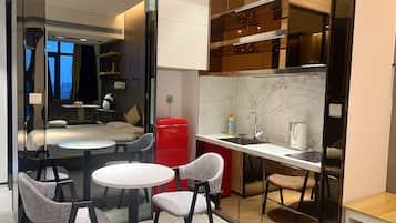 A compact hotel room with a bed, a small dining table, two chairs, a red cabinet, a kitchen area with a sink and stove, and a large mirror.