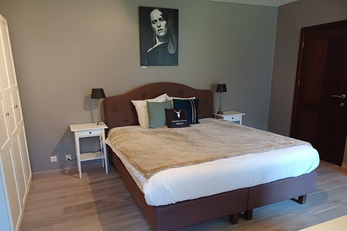 A bedroom with a large bed, two bedside tables, a nightstand, a wall-mounted painting, and a wooden floor.