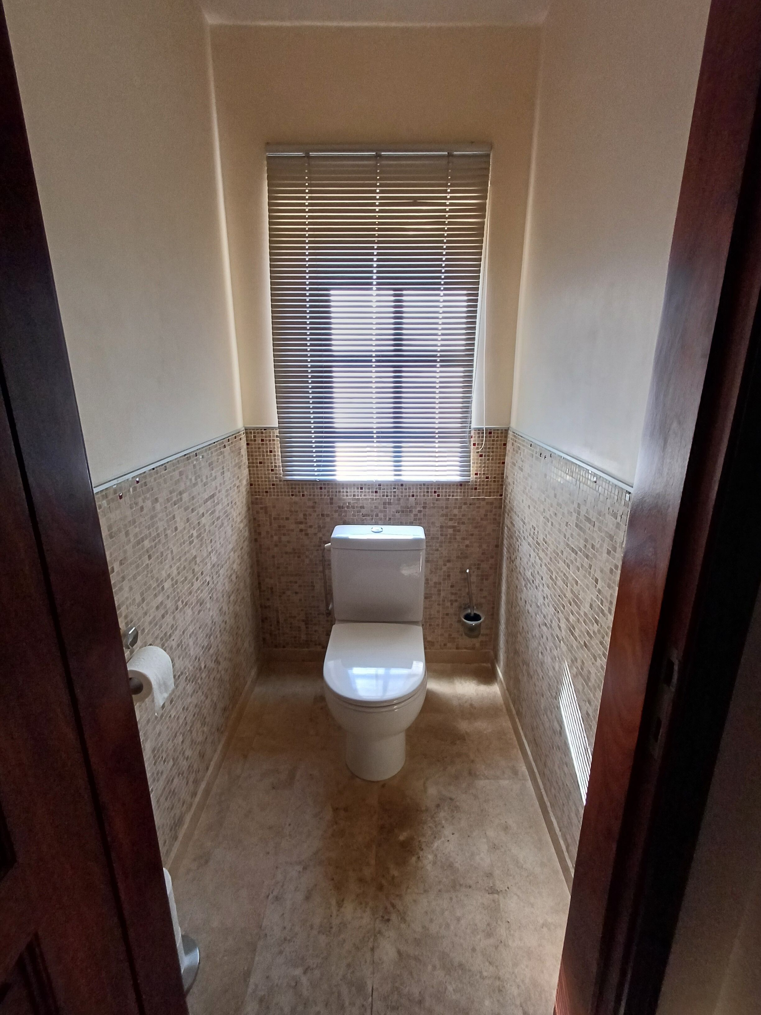 A small bathroom with a toilet, a window with blinds, and a tiled wall.