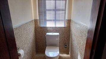 A small bathroom with a toilet, a window with blinds, and a tiled wall.