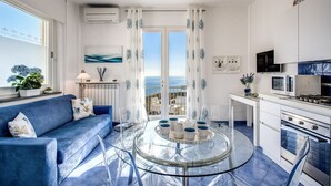 A modern kitchen with a glass dining table, a blue sofa, and a view of the sea through large windows.