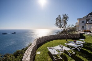 A coastal property with a stone retaining wall, a grassy lawn, and lounge chairs facing the sea.