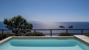 A clear view of a swimming pool with a wooden deck and a railing, overlooking a calm sea with distant islands.