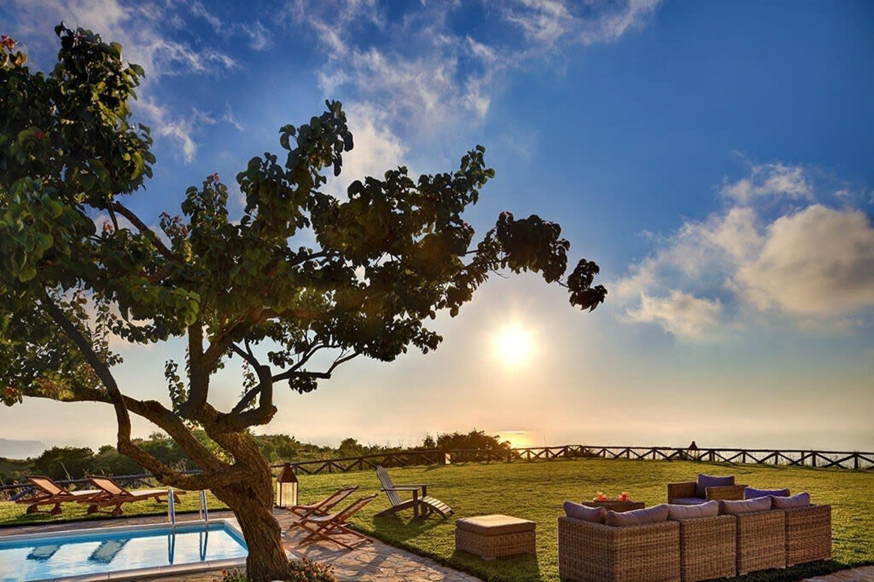Área da piscina com espreguiçadeiras e um sofá de vime, cercada por um gramado e uma árvore.