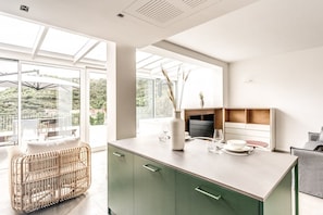 A modern kitchen with a large island, green cabinets, and a view of a garden through large windows.