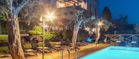 A swimming pool with lounge chairs and a hot tub, surrounded by trees and a building with illuminated windows.