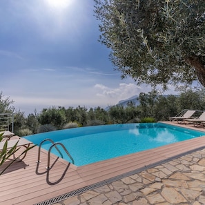 A clear blue swimming pool with a wooden deck and lounge chairs, surrounded by greenery and a mountain view.