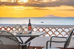 Ein Tisch mit einer Flasche, Gläsern und einem Buch auf einem Balkon mit Blick auf das Meer und die Berge.