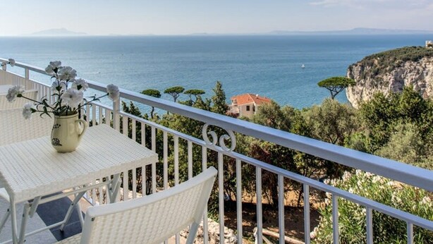 A balcony with a white table and chair, overlooking a coastal view with a clear sky and distant land. - Casa Tit in Massa Lubrense (Massa Lubrense)