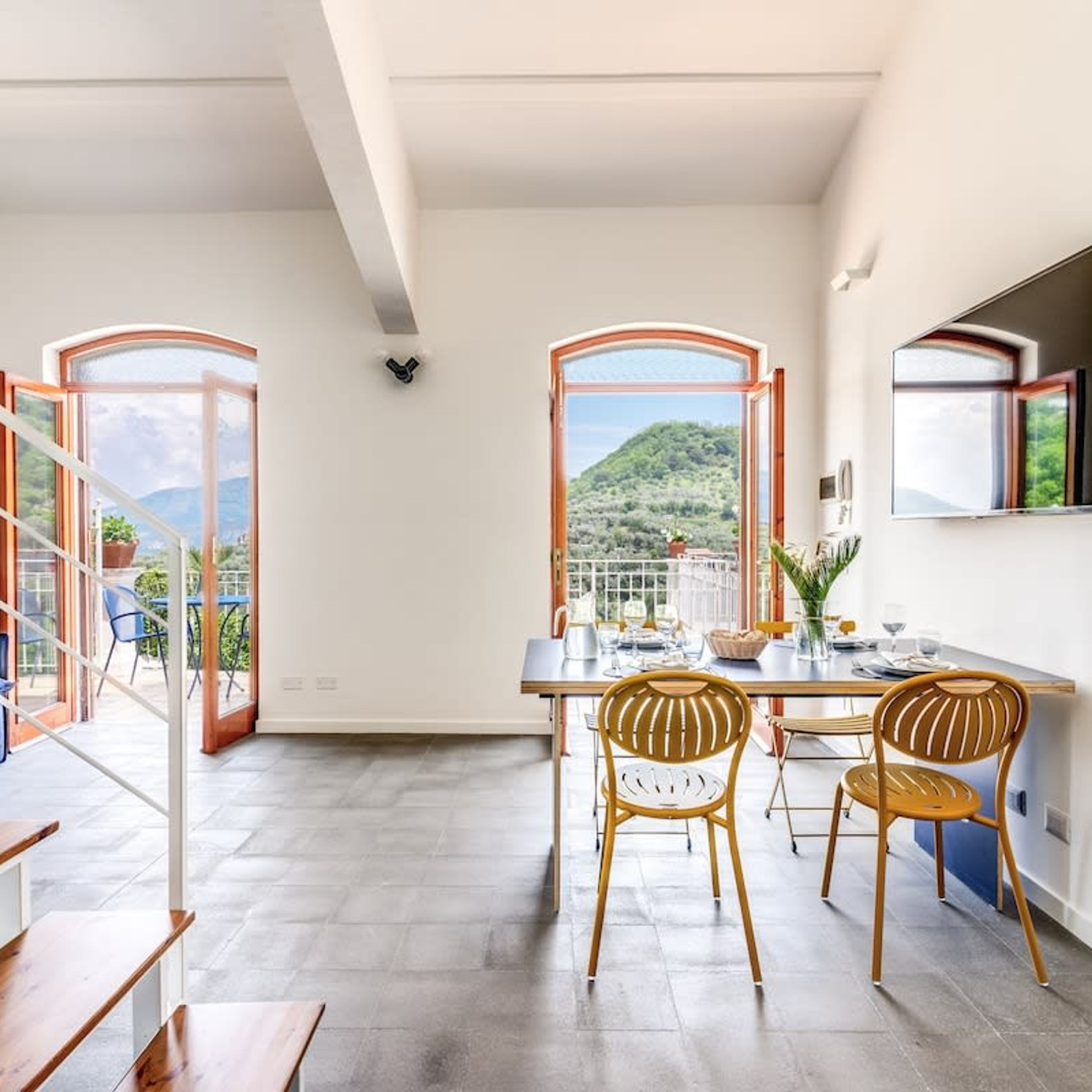 Comedor con una mesa dispuesta para cuatro, dos sillas de mimbre y vistas a un paisaje montañoso a través de ventanas amplias.
