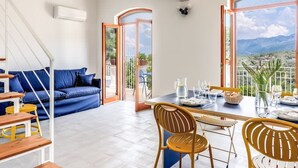 Un vaste espace de vie comprenant un canapé bleu, une table à manger en bois et des chaises, de grandes fenêtres offrant une vue sur les montagnes, et un escalier menant à un autre niveau.
