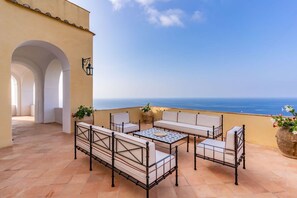 Eine Terrasse mit Gartenmöbeln, Blick auf das Meer und ein Gebäude mit Bögen.