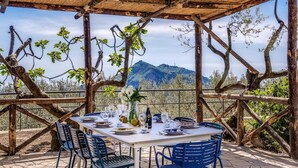 An outdoor dining area with a table set for four, surrounded by chairs, under a wooden pergola with a view of mountains. - Villa Rusinella in Massa Lubrense (Massa Lubrense)