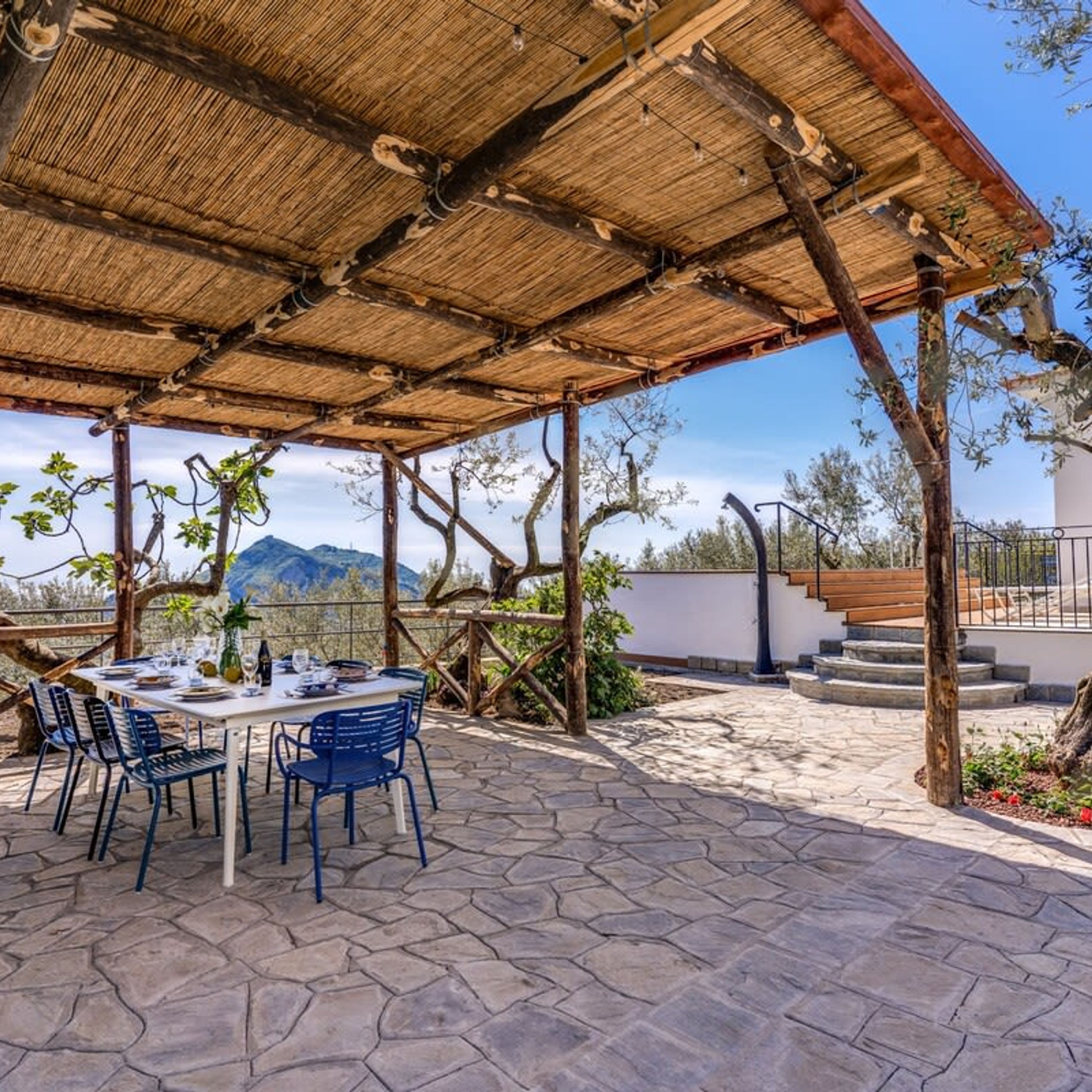 An outdoor patio with stone flooring, a covered seating area with tables and chairs, and a view of mountains in the background.