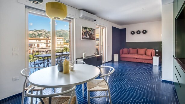 Una sala de estar moderna con piso de espiga azul, una mesa redonda blanca con sillas, un sofá rosa y un ventanal con vistas a edificios y colinas.