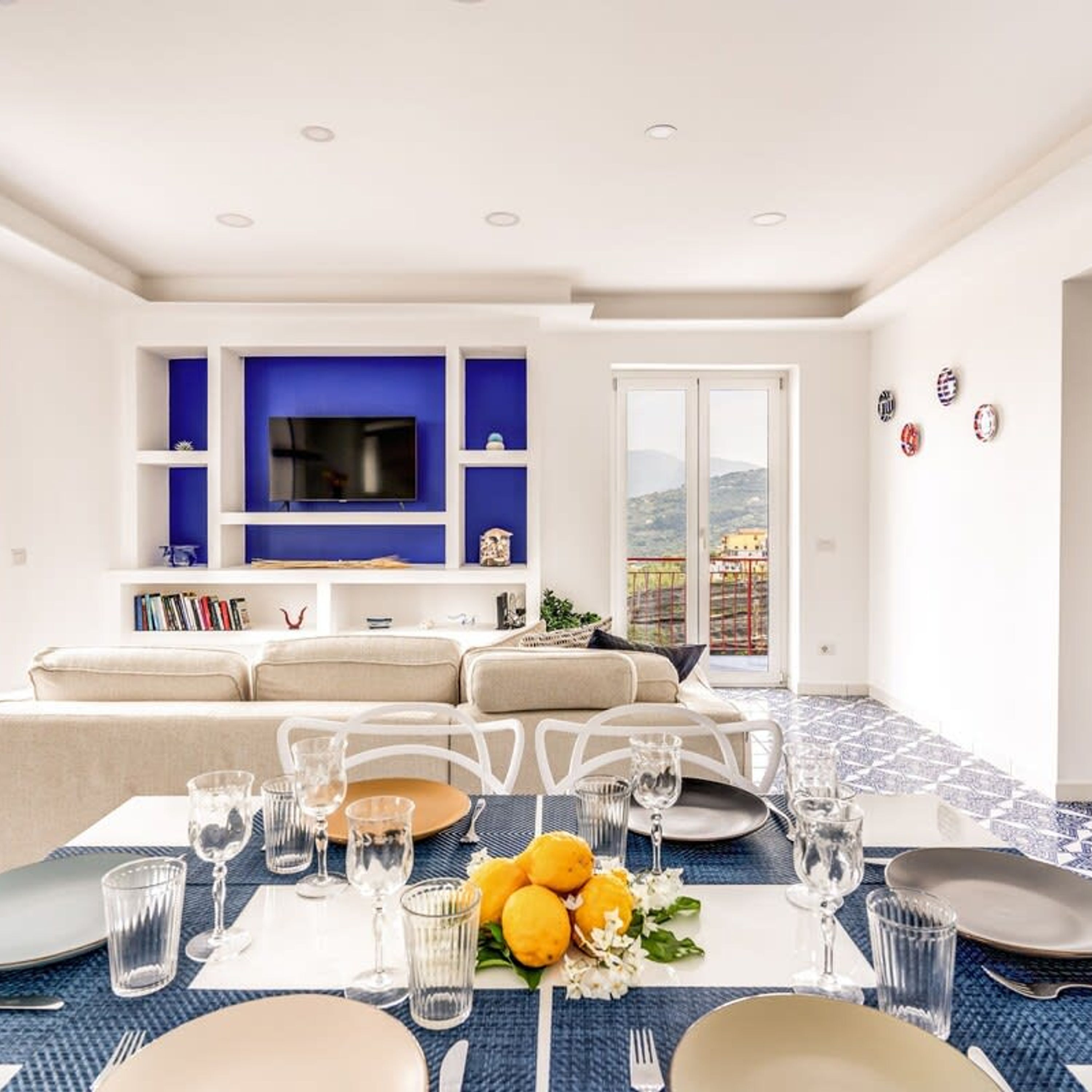 A modern living room with a flat-screen TV, blue shelving, a sofa, and a dining area set for four.