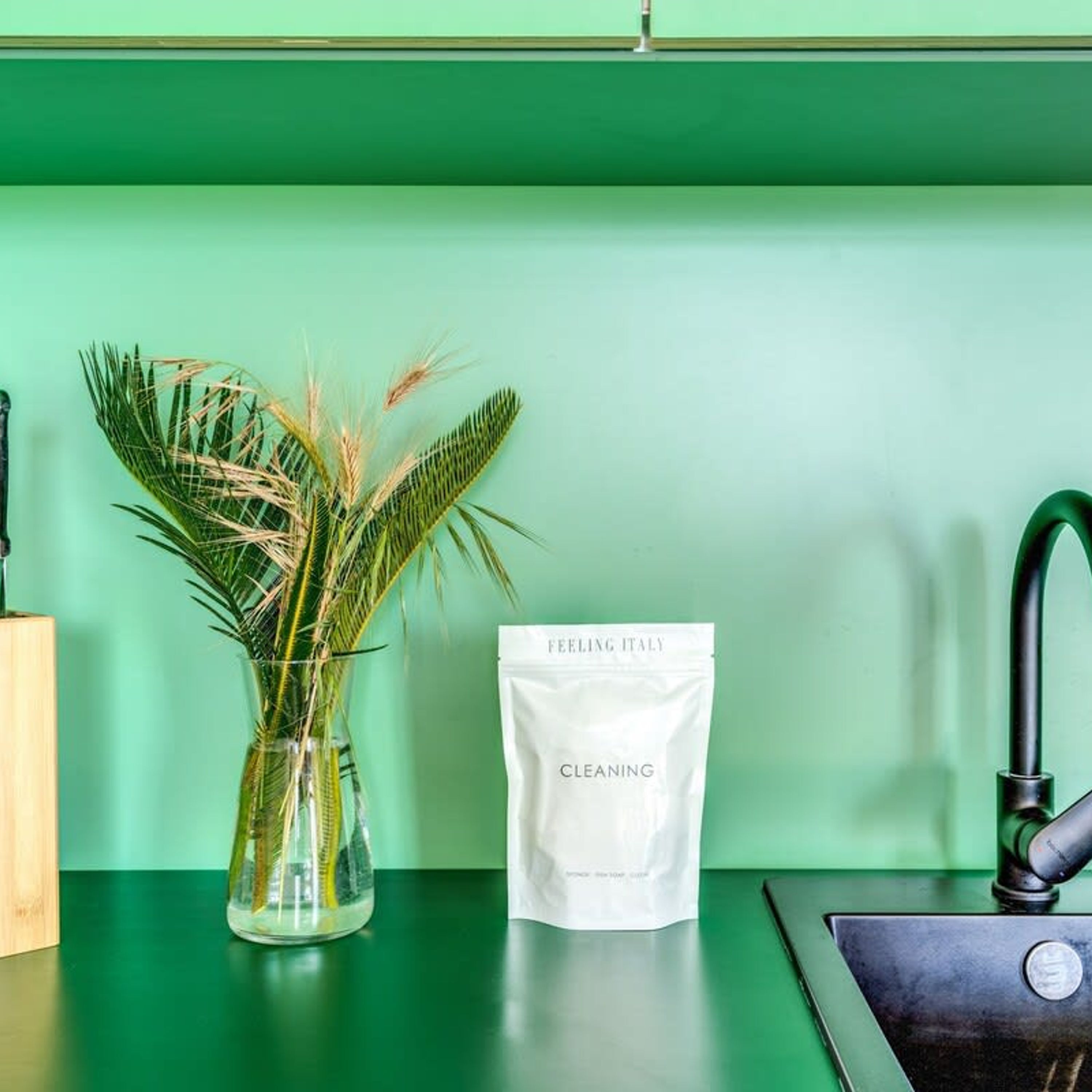 A kitchen counter with a green wall, a clear glass vase with dried plants, and a white bag labeled "FEELING ITALY CLEANING".
