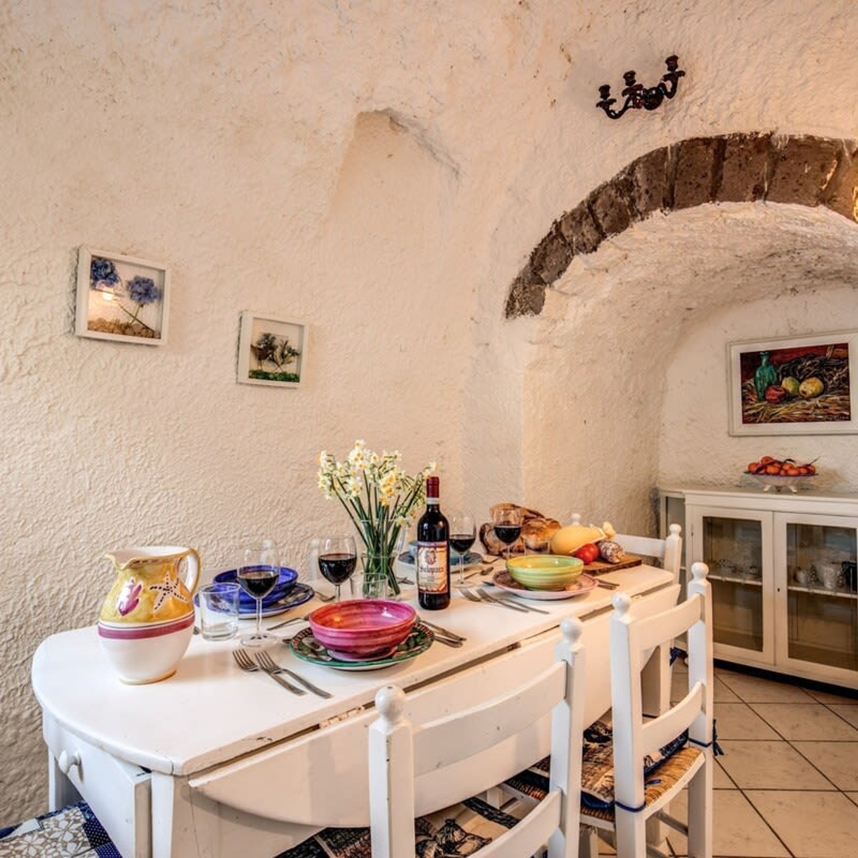 A dining area with a rustic stone wall, a white table set with plates, glasses, and a bottle, and a cabinet with glass doors in the background.