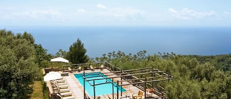 A stone pool with a wooden deck, surrounded by lush greenery and a view of the sea.