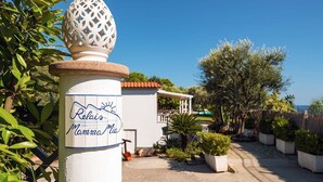 Une enseigne portant l’inscription « Relais Mamma Mia », un bâtiment blanc avec un toit recouvert de tuiles rouges, et un jardin agrémenté de plantes en pot et d’une pergola.