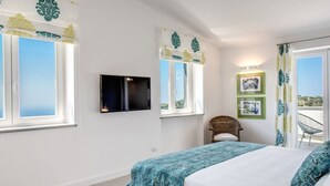 Ein Schlafzimmer mit einem Bett, einem Sessel, einem Fernseher und zwei mit Vorhängen versehenen Fenstern.
