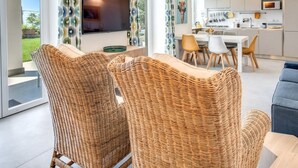 Ein modernes Wohnzimmer mit Rattanmöbeln, einem Flachbildfernseher und einem Essbereich, im Hintergrund ist eine Küche zu sehen.