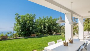 Ein moderner Essbereich im Freien mit einem weißen Tisch und Stühlen, einer hängenden Pendelleuchte und Blick auf einen üppigen Garten und das Meer.