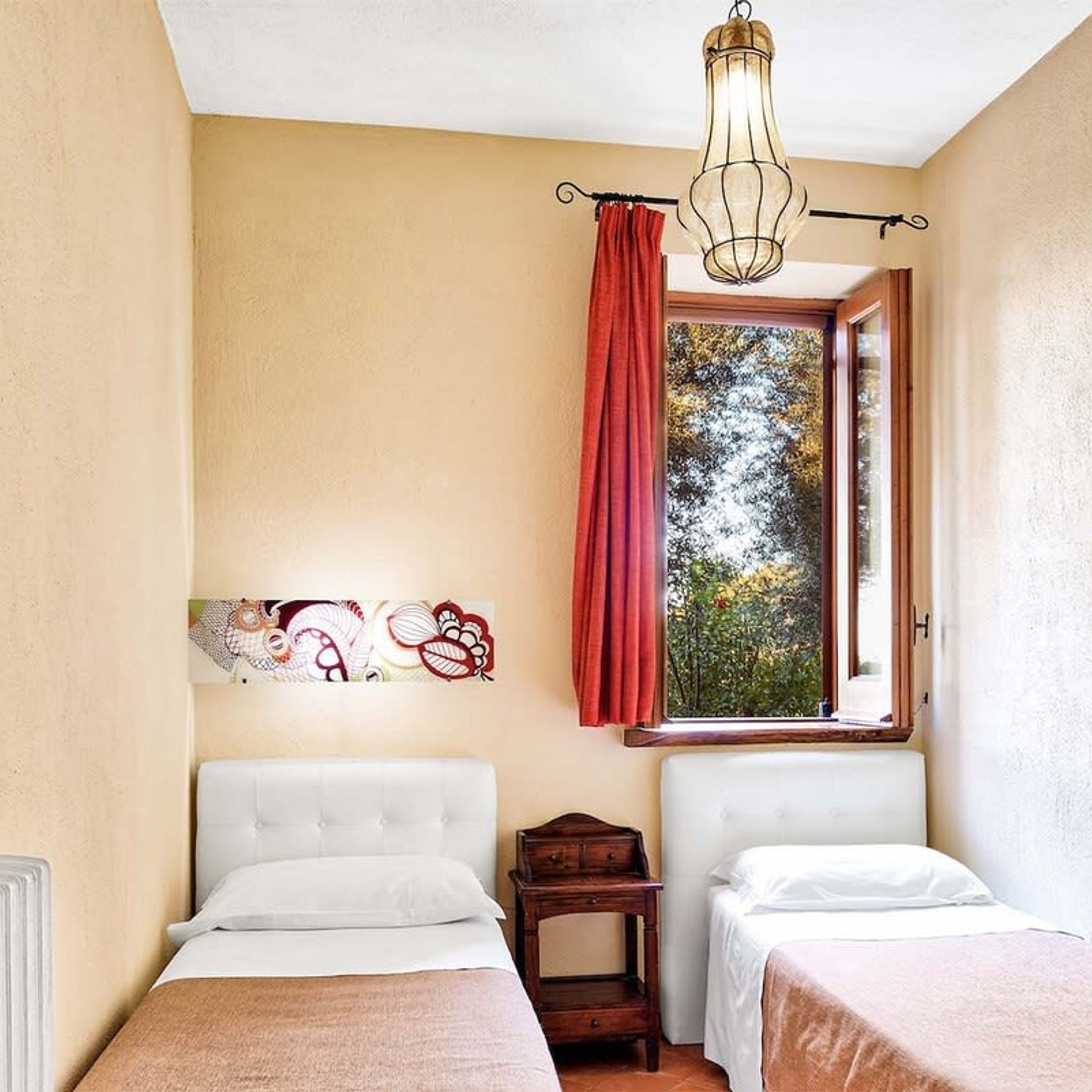 Un dormitorio con dos camas, una ventana que da a árboles, una lámpara colgante y una mesita de noche de madera.