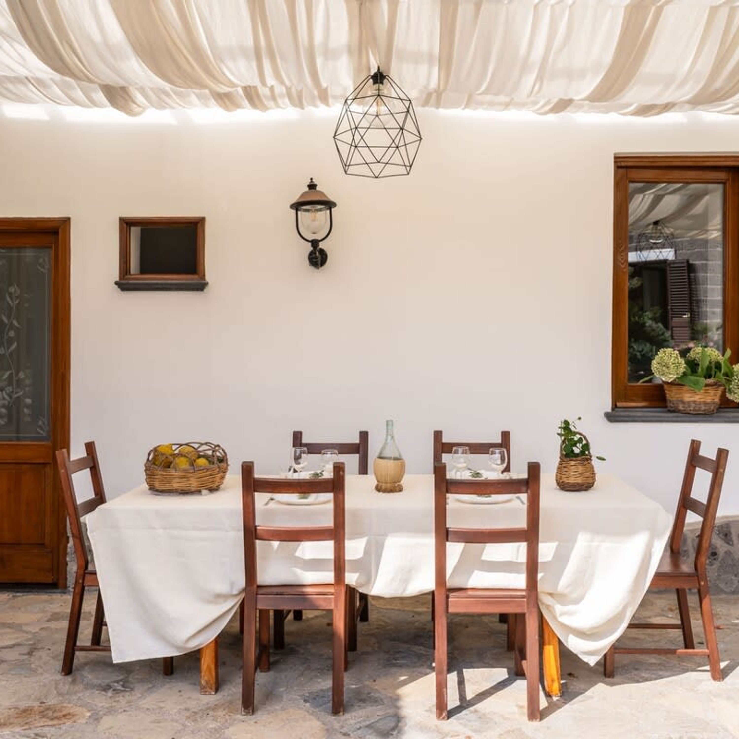 Un espace extérieur abrité, aménagé pour les repas, avec une table dressée pour quatre personnes, entourée de chaises en bois, et un luminaire suspendu au-dessus.