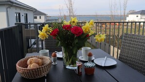 A table set with bread, eggs, a jar of jam, and a vase of flowers on a balcony overlooking a body of water.