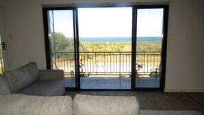 A living room with a large sliding glass door leading to a balcony with a view of the sea and greenery.