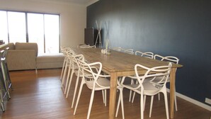 A dining area with a long wooden table surrounded by white chairs, a sofa, and a TV against a dark wall.