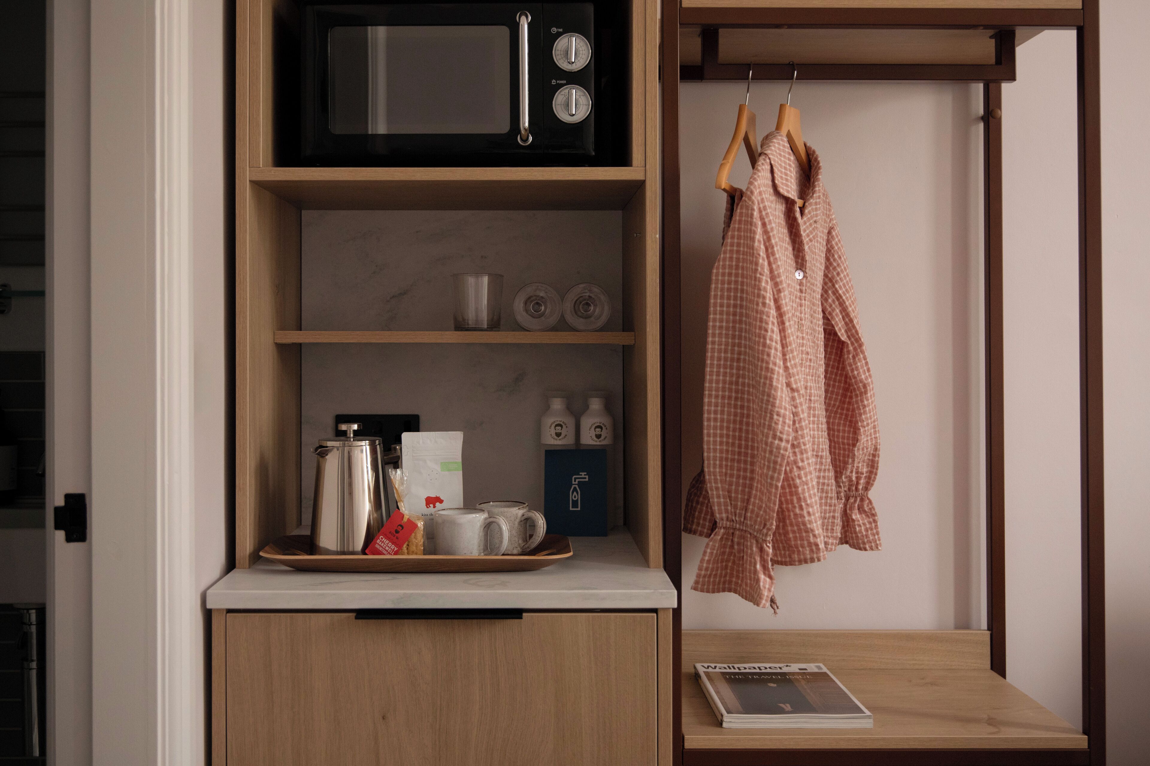 A kitchenette with a microwave, coffee maker, and tea set. There is a shelf with bottles and a tray with a teapot and cups. A checkered shirt hangs on a hanger to the right.
