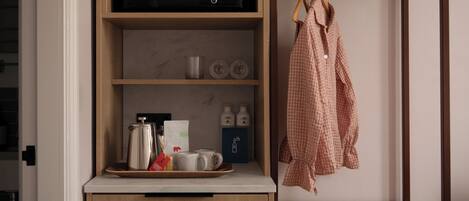 A kitchenette with a microwave, coffee maker, and tea set. There is a shelf with bottles and a tray with a teapot and cups. A checkered shirt hangs on a hanger to the right.