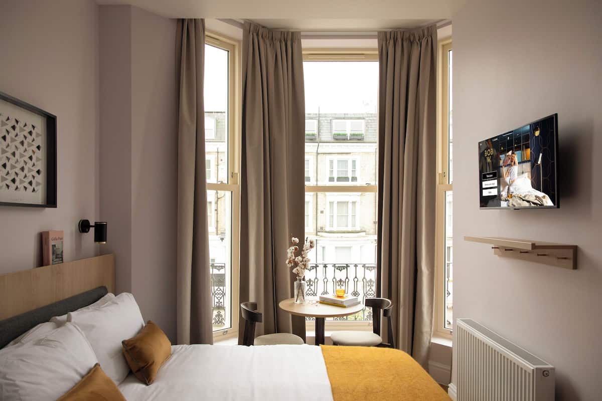 Een hotelkamer met een bed, een aan de muur bevestigde televisie, een raam met gordijnen, een klein tafeltje met een vaas en een boek, en een radiator.