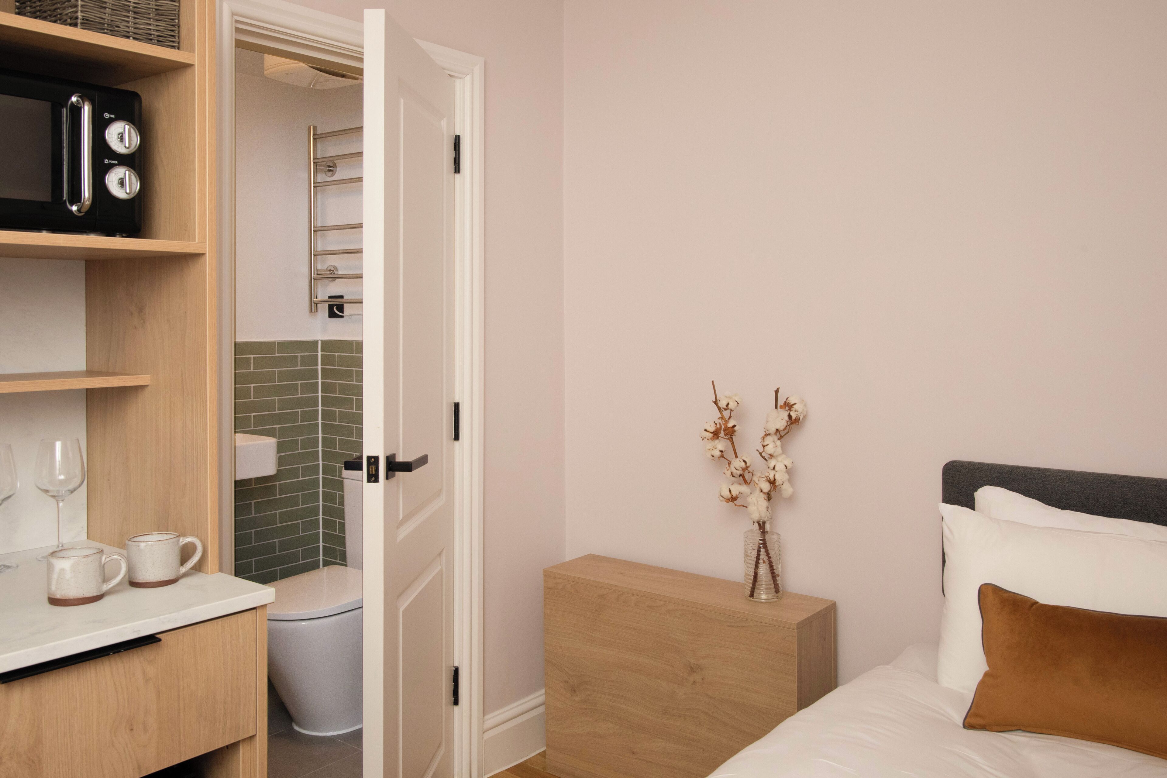 Ein Hotelzimmer mit einem Holzschrank, einem Nachttisch mit Blumenvase, einem Bett mit weißer Bettwäsche und einem braunen Kopfkissen sowie einer Mikrowelle auf einem Regal.