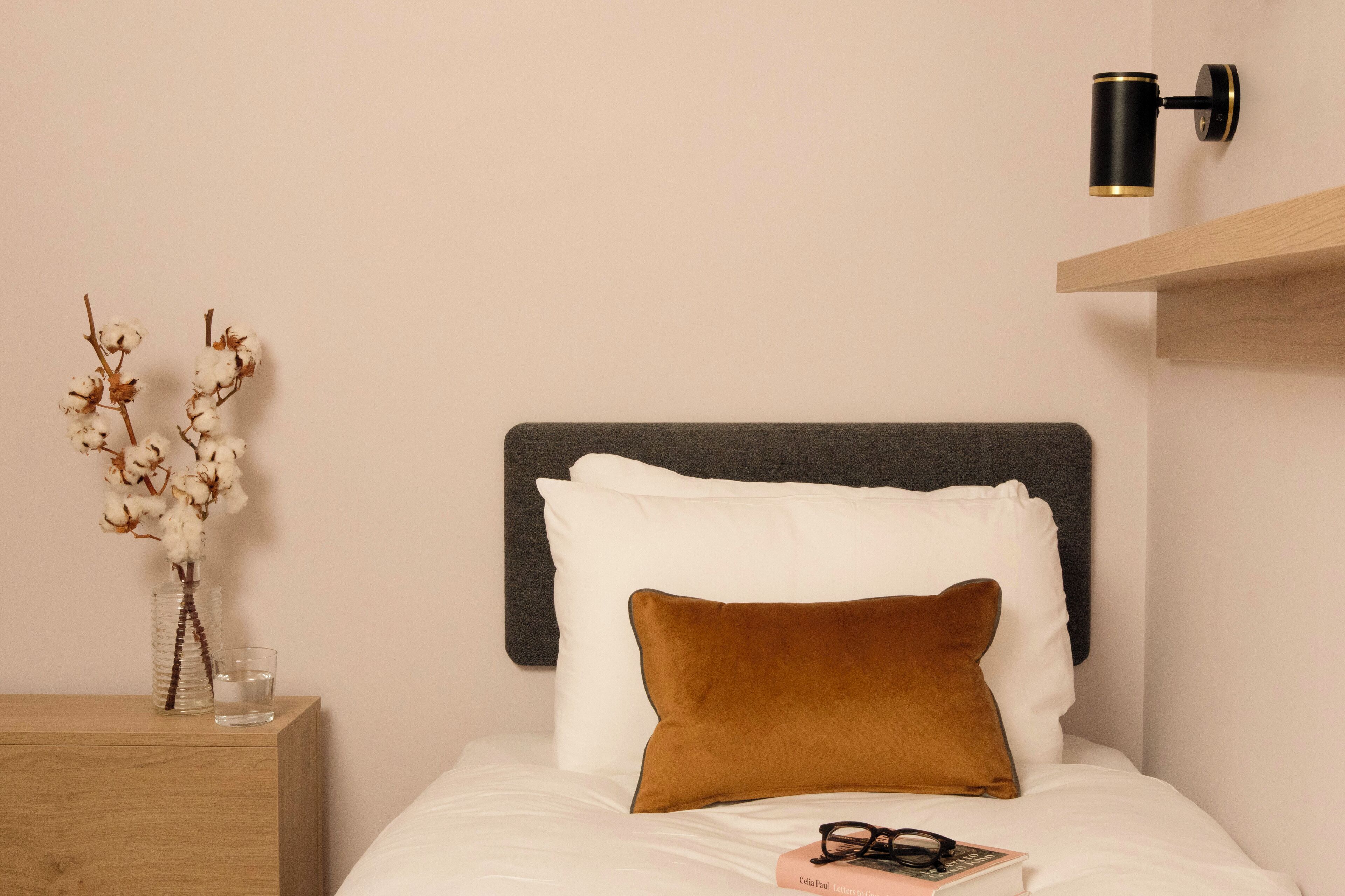 A neatly made bed with a dark headboard, a brown pillow, and a pair of glasses on the bed. There is a small vase with dried flowers on a wooden nightstand beside the bed.